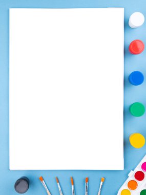 Üzerinde fırça, boya kalemleri, suluboya ve diğer kırtasiye malzemelerinin bulunduğu boş bir defterin beyaz sayfası olan düz bir ofis masası. Üst manzara, düz uzanma.