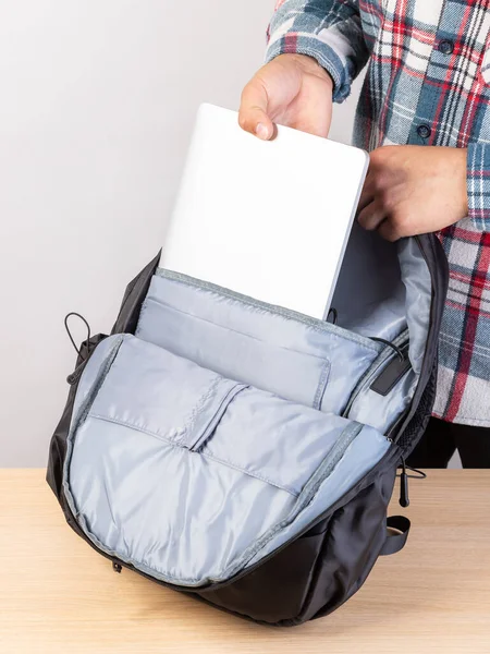 A man puts a laptop in a backpack outside, on a light background. A young man packs a laptop into a backpack. A man pulls a laptop out of his backpack.