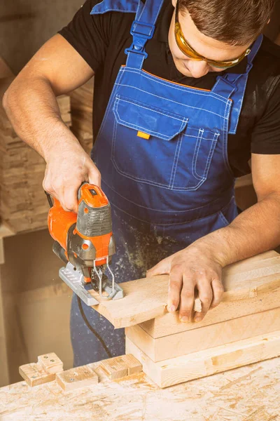 Close up of experienced carpenter in work clothes and small buiness owner  carpenter saw and processes the edges of a wooden bar with a jig saw  in a workshop
