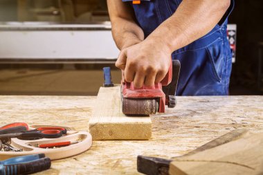 İş kıyafetleri konusunda deneyimli marangoz ve ahşap atölyesinde çalışan küçük büro sahibi atölyede ahşap cilalamak için zımpara kağıdı kullanıyor.  