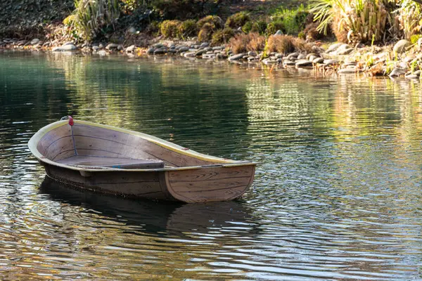A small wooden rowing boat floats on calm waters with reeds in the water. Reflections of the boat are visible in the water.