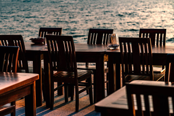 Row of wooden tables and chairs of a restaurant on the beach of Thailand, no people, sunset time, blurred background of sea wave.
