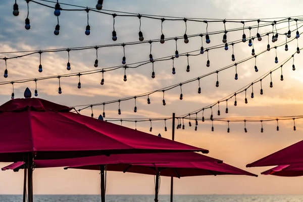 Birçok plaj kırmızı şemsiyesinin üst kısmı ve birçok parti lambası havada asılı, denizin üzerinde renkli turuncu gökyüzü.
