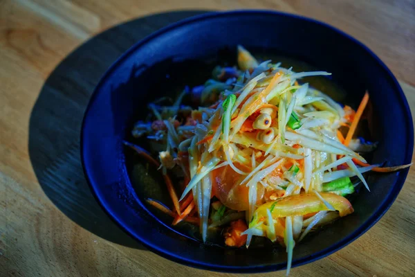 Thai food called Som Tam or spicy and sour fresh papaya salad, food in black food plate on wooden table, natural sunlight.