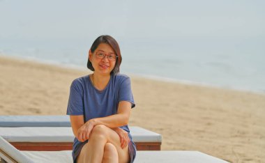 Orta yaşlı Asyalı kadın deniz kenarında güneşli bir yatakta oturuyor. Sahilde güneş gözlüğü takan sağlıklı Asyalı kadın. Gün ışığı, mesaj ve tasarım için boş alan..