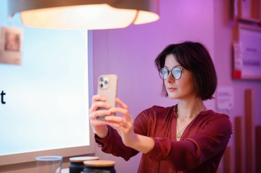 Gözlüklü şık bir kadın akıllı telefonuyla selfie çekerken saçını düzeltir. Sıcak ortam aydınlatması ve modern bir iç mekan rahat ve modaya uygun bir atmosfer yaratır..