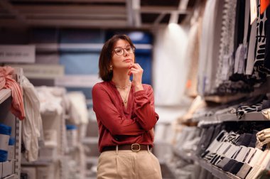 Gözlüklü şık bir kadın bir tekstil mağazasında durup seçeneklerini değerlendiriyor. Modern perakende satışı moda ve zarif bir alışveriş deneyimi yaratıyor..