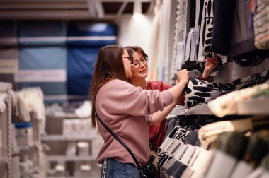 Gülümseyen iki kadın ev dokusunda geziniyor, keyifli bir alışveriş deneyimi paylaşıyorlar. Modern bir mağazada kumaşı birlikte incelerler, ev dekoru seçerken eğlenirler..