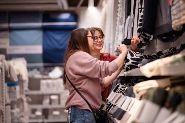 Gülümseyen iki kadın ev dokusunda geziniyor, keyifli bir alışveriş deneyimi paylaşıyorlar. Modern bir mağazada kumaşı birlikte incelerler, ev dekoru seçerken eğlenirler..