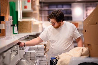 Beyaz tişörtlü ve gözlüklü bir adam bir mağaza koridorunda ev eşyalarına göz atıyor, raftan bir ürün seçerken ev malzemeleriyle dolu bir arabayı itiyor..