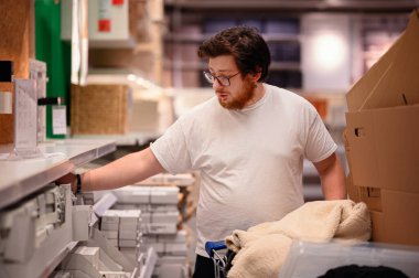 Beyaz tişörtlü ve gözlüklü bir adam bir mağaza koridorunda ev eşyalarına göz atıyor, raftan bir ürün seçerken ev malzemeleriyle dolu bir arabayı itiyor..