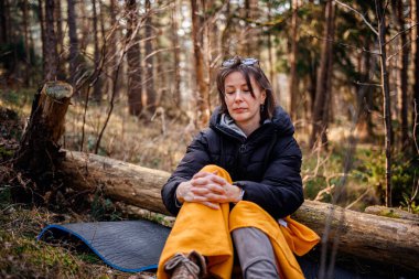 Siyah ceketli ve sarı battaniyeli bir kadın ormanda bir kütüğe oturmuş, elleri sımsıkı bağlanmış, doğal ortamda düşünceli ve huzurlu görünüyor..