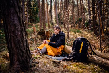 Siyah ceketli ve sarı battaniyeli bir kadın ormanda bir kütüğe oturur, elleri sımsıkı bağlanmış, ağaçlar ve sırt çantasıyla çevrili, doğada sakin bir anın tadını çıkarır..