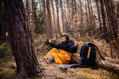 Siyah ceketli ve sarı battaniyeli bir kadın bir kütüğün üzerinde oturuyor, kulaklık ve güneş gözlüğü takıyor, huzurlu bir ormanda müziğe neşeyle tepki veriyor..