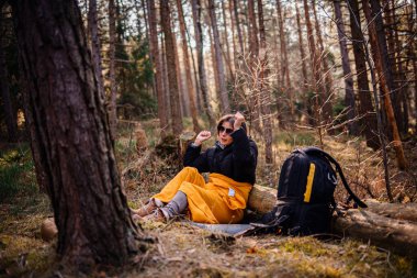 Siyah ceketli ve sarı battaniyeli bir kadın bir kütüğün üzerinde oturuyor, kulaklık ve güneş gözlüğü takıyor, huzurlu bir ormanda müziğe neşeyle tepki veriyor..