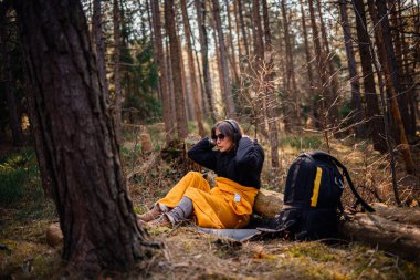 Siyah ceketli ve sarı battaniyeli bir kadın ormanda bir kütüğe oturmuş, kulaklıklarını düzeltirken doğanın tadını çıkarıyor. Yakınlarda bir sırt çantası duruyor..