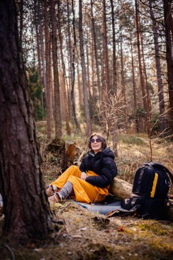 Siyah ceketli ve sarı battaniyeli bir kadın ormanda bir kütüğe yaslanır, güneş gözlüğü takar ve huzurlu doğal çevrenin tadını çıkarır. Yakınlarda bir sırt çantası var..