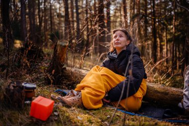 Siyah ceketli ve sarı battaniyeli bir kadın ormanda bir paspasın üzerinde oturur, düşünceli bir ifadeyle yukarıya bakar, kamp malzemeleri ve doğal manzarayla çevrilidir..