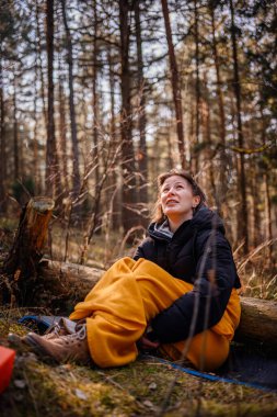 Siyah ceketli ve sarı battaniyeli bir kadın ormanda bir paspasın üzerinde oturur, düşünceli bir ifadeyle yukarıya bakar, kamp malzemeleri ve doğal manzarayla çevrilidir..