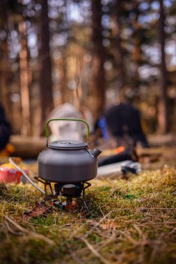 Orman ortamında taşınabilir bir ocakta yeşil saplı metal bir çaydanlık, kamp malzemeleri ve arka planda çadırlarla çevrili..