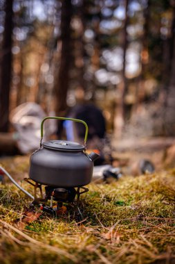 Orman ortamında taşınabilir bir ocakta yeşil saplı metal bir çaydanlık, kamp malzemeleri ve arka planda çadırlarla çevrili..