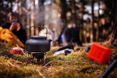 Yeşil saplı buharlı bir metal çaydanlık ormandaki portatif bir sobanın üzerinde oturur. Arka planda bir karavan kamp malzemelerinin arasında sıcak bir içeceğin keyfini çıkarır..