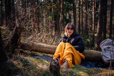 Siyah ceketli ve sarı battaniyeli bir kadın güneşli bir ormanda bir kütüğe oturur, güneş gözlüğü ve botlar takar, yanında bir sırt çantasıyla, huzurlu bir şekilde dışarıda olmanın tadını çıkarır..