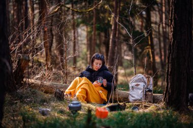 Siyah ceketli ve sarı battaniyeli bir kadın ormandaki bir kütüğe oturur, elinde bir bardak, kamp malzemeleri ve sırt çantasıyla, huzurlu bir şekilde dışarıda oturmanın tadını çıkarır..