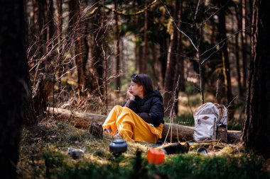 Siyah ceketli ve sarı battaniyeli bir kadın ormanda bir kütüğün üzerinde oturuyor, uzaklığa bakıyor, sırt çantası ve kamp malzemeleriyle çevrili..