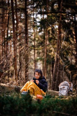 Siyah ceketli ve sarı battaniyeli bir kadın ormanda bir kütüğün üzerinde oturuyor, uzaklığa bakıyor, sırt çantası ve kamp malzemeleriyle çevrili..