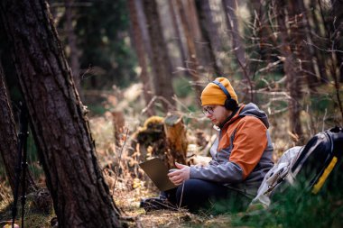 Bere ve kulaklıklı bir adam ormanda yerde oturuyor, dizüstü bilgisayarına odaklanmış, sırt çantası, fincan ve açık hava teçhizatı ile çevrili, doğada uzaktan çalışmanın keyfini çıkarıyor..