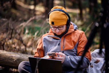 Bere ve kulaklık takmış bir adam ormanda yerde oturuyor, akıllı telefonunu laptopta çalışırken kullanıyor, sırt çantası, tripod ve kamp malzemeleriyle çevrili..