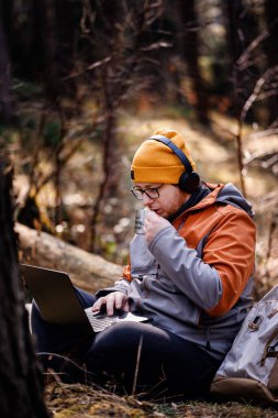 Bere ve kulaklıklı bir adam ormanda oturuyor, elinde kahve fincanı, sırt çantası ve açık hava teçhizatıyla dizüstü bilgisayarıyla çalışıyor..
