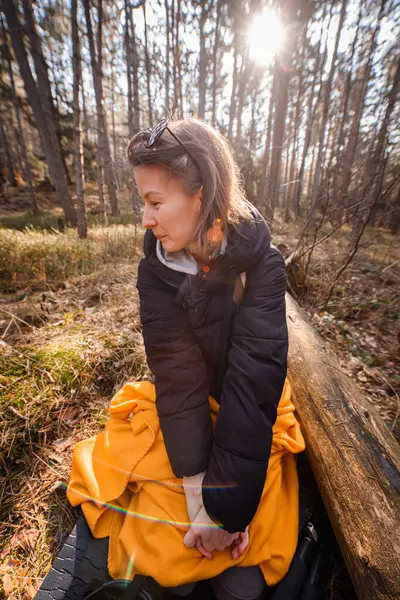 Siyah ceketli genç bir kadın ormanda bir kütüğe oturmuş, sarı bir battaniyeye sarılmış, ağaçların arasından süzülen güneş ışığını seyrediyor..