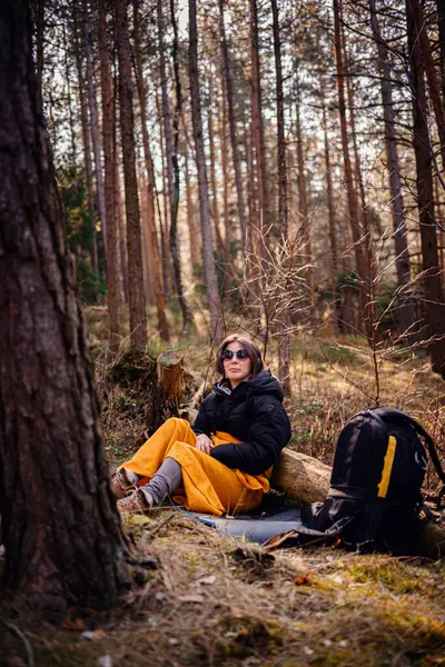 Siyah ceketli ve sarı battaniyeli bir kadın ormanda bir kütüğe yaslanır, güneş gözlüğü takar ve huzurlu doğal çevrenin tadını çıkarır. Yakınlarda bir sırt çantası var..