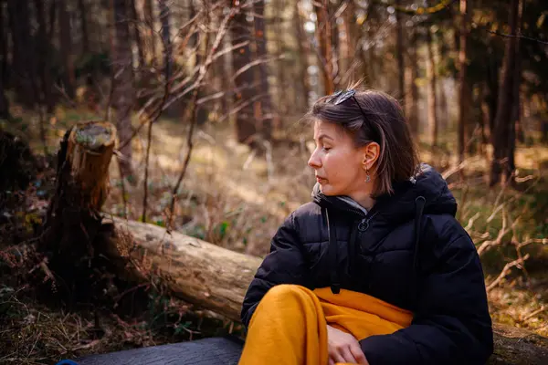 Siyah ceketli ve sarı battaniyeli bir kadın ormanda bir paspasın üzerinde oturur, ağaçlar ve güneş ışığıyla çevrili, huzurlu bir ifadeyle yan tarafa bakar..
