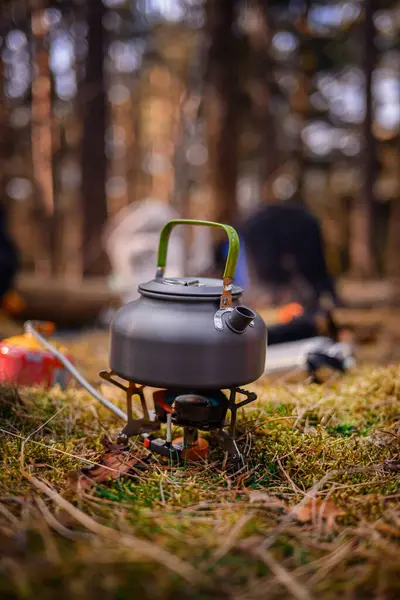 Orman ortamında taşınabilir bir ocakta yeşil saplı metal bir çaydanlık, kamp malzemeleri ve arka planda çadırlarla çevrili..