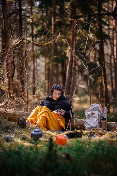 Siyah ceketli ve sarı battaniyeli bir kadın ormandaki bir kütüğe oturur, elinde bir bardak, kamp malzemeleri ve sırt çantasıyla, huzurlu bir şekilde dışarıda oturmanın tadını çıkarır..