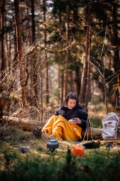 Siyah ceketli ve sarı battaniyeli bir kadın ormandaki bir kütüğe oturur, elinde bir bardak, kamp malzemeleri ve sırt çantasıyla, huzurlu bir şekilde dışarıda oturmanın tadını çıkarır..