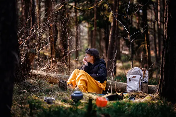 Siyah ceketli ve sarı battaniyeli bir kadın ormanda bir kütüğün üzerinde oturuyor, uzaklığa bakıyor, sırt çantası ve kamp malzemeleriyle çevrili..