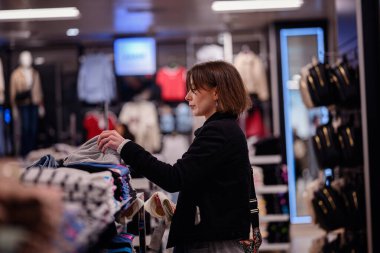 Siyah ceketli şık bir kadın bir moda mağazasında katlanmış kıyafetleri karıştırıyor ve dolabı için dikkatlice seçiyor..