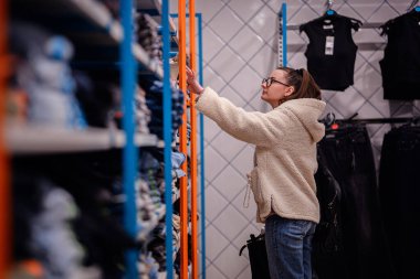 Rahat yün ceketli bir kadın mağazadaki raflarda katlanmış kıyafetlere göz atıyor, moda bir mağazada alışveriş yaparken bir eşyaya uzanıyor..