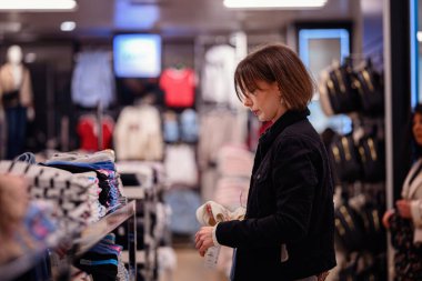 Siyah ceketli şık bir kadın bir moda mağazasında katlanmış kıyafetleri karıştırıyor ve dolabı için dikkatlice seçiyor..