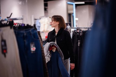 Siyah ceketli bir kadın, bir moda mağazasında rafları karıştırırken, kıyafetlerini seçerken birden fazla giysi taşır..