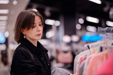 Siyah ceketli şık bir kadın giyim mağazasında yumuşak, renkli kazaklara göz gezdiriyor ve gardırobu için dikkatlice seçiyor..