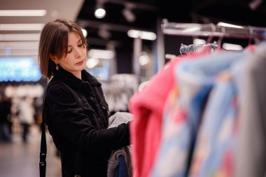 Siyah ceketli şık bir kadın giyim mağazasında yumuşak, renkli kazaklara göz gezdiriyor ve gardırobu için dikkatlice seçiyor..