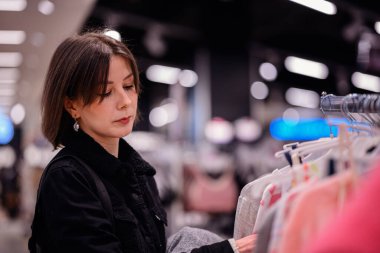 Siyah ceketli şık bir kadın giyim mağazasında yumuşak, renkli kazaklara göz gezdiriyor ve gardırobu için dikkatlice seçiyor..