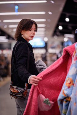 Siyah ceketli şık bir kadın giyim mağazasında yumuşak, renkli kazaklara göz gezdiriyor ve gardırobu için dikkatlice seçiyor..