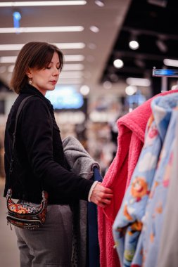 Siyah ceketli şık bir kadın giyim mağazasında yumuşak, renkli kazaklara göz gezdiriyor ve gardırobu için dikkatlice seçiyor..
