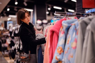 Siyah ceketli şık bir kadın giyim mağazasında yumuşak, renkli kazaklara göz gezdiriyor ve gardırobu için dikkatlice seçiyor..